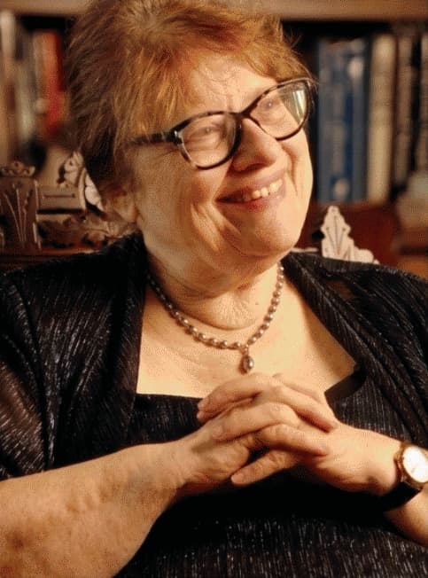 Janice Stauffer smiling, hands clasped, seated in front of a bookshelf